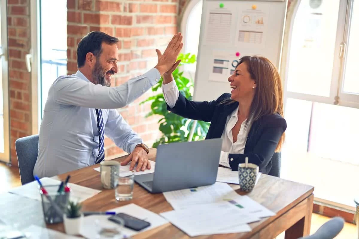 Deux professionnels se félicitent en se tapant dans la main autour d’un bureau, devant un ordinateur portable et des Deux professionnels se félicitent en se tapant dans la main autour d’un bureau, devant un ordinateur portable et des