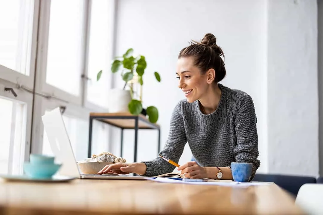 Femme travaillant sur un ordinateur portable à domicile, souriante, avec carnet et stylo sur la table, ambiance lumineuse et Femme travaillant sur un ordinateur portable à domicile, souriante, avec carnet et stylo sur la table, ambiance lumineuse et